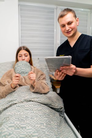 Foto de El médico en la habitación del paciente después de la cirugía plástica sostiene una tableta en sus manos la chica se mira en el espejo y evalúa el resultado - Imagen libre de derechos