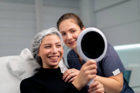 Photo for Satisfied beautiful female client in the beauty salon looks in the mirror together with the doctor checks the result of cosmetic procedure - Royalty Free Image