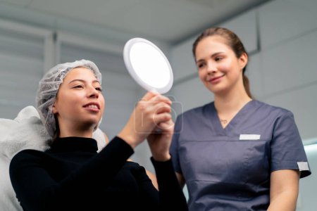 Photo for Satisfied beautiful female client in the beauty salon looks in the mirror together with the doctor checks the result of cosmetic procedure - Royalty Free Image