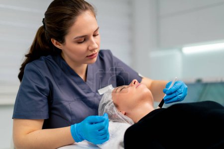 Foto de Médico esteticista en guantes sosteniendo un cepillo y aplicando una máscara de salud en la cara y el cuello de una cliente femenina en el salón de belleza - Imagen libre de derechos