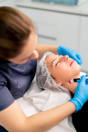 Téléchargez les photos : Esthéticienne médecin en gants tenant une brosse et appliquant un masque de santé sur le visage et le cou d'une cliente dans un salon de beauté - en image libre de droit