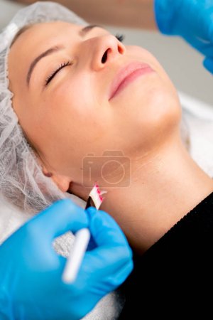 Foto de Primer plano de las manos del médico esteticista en guantes sosteniendo un cepillo y aplicando una máscara de salud en la cara y el cuello de una clienta en el salón de belleza - Imagen libre de derechos