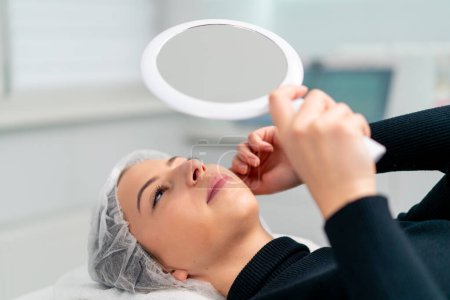 Foto de La cara satisfecha de una clienta en un salón de belleza se mira en el espejo y disfruta del resultado de un procedimiento cosmético - Imagen libre de derechos