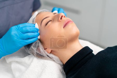 Téléchargez les photos : Gros plan un cosmétologue nettoie la peau d'un client avec un coton-tige avant de commencer la procédure de beauté et de santé - en image libre de droit