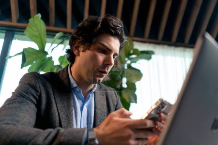 Téléchargez les photos : Portrait d'un homme d'affaires sérieux travaillant sur un ordinateur portable au bureau vérifiant la chute du marché boursier avec smartphone - en image libre de droit