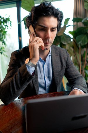 Foto de Retrato de un hombre serio un hombre de negocios trabaja en una computadora portátil en la oficina se comunica con sus colegas en el teléfono inteligente - Imagen libre de derechos