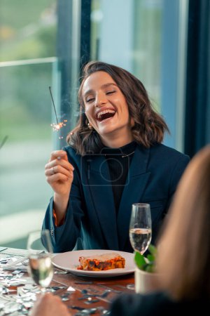 Foto de Retrato de una hermosa mujer sonriente sosteniendo un bengala durante una celebración de cumpleaños en un restaurante en la compañía de amigos - Imagen libre de derechos