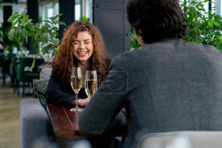 Téléchargez les photos : Portrait d'une jeune femme bouclée souriante lors d'une réunion d'affaires avec un collègue ou lors d'un rendez-vous avec un homme avec des verres de champagne célébration - en image libre de droit