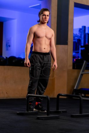 Photo for In a sports club a guy trainer in black sweatpants stands near the floor horizontal bar - Royalty Free Image