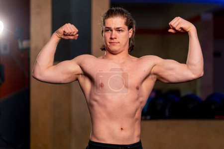 Photo for In a sports club guy trainer in black sweatpants stands and demonstrates the muscles on his arms - Royalty Free Image