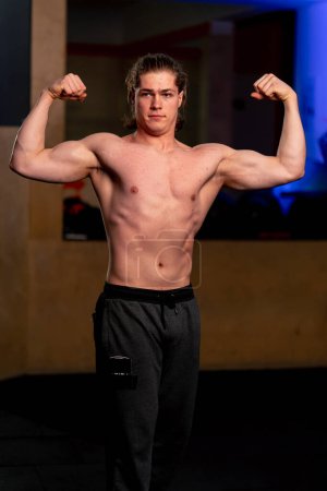 Photo for In a sports club guy trainer in black sweatpants stands and demonstrates the muscles on his arms - Royalty Free Image