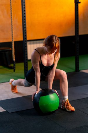Photo for In a sports club red-haired trainer does a warm-up for her legs leaning on a ball - Royalty Free Image