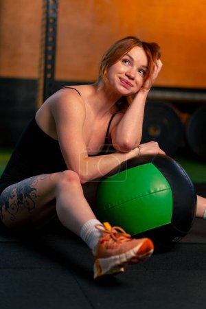Photo for In the gym a coach girl sits on the floor hugging a large bench press ball - Royalty Free Image