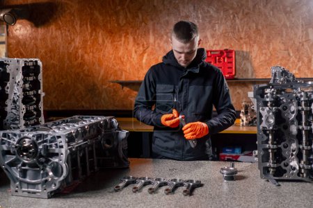 Photo for At the service station on the table a young engine repairman examines a new part for the piston - Royalty Free Image