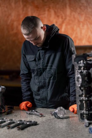 Foto de En una estación de servicio sobre la mesa un joven reparador de motores monta una nueva pieza de pistón - Imagen libre de derechos