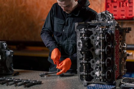 Foto de En una estación de servicio sobre la mesa un joven reparador de motores monta una nueva pieza de pistón - Imagen libre de derechos