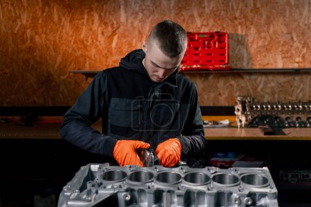Foto de En una estación de servicio en la mesa un joven reparador de motores pone una pieza de pistón en su lugar - Imagen libre de derechos