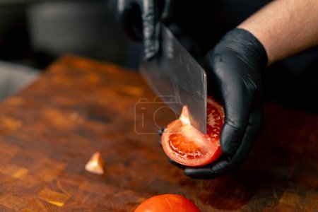 Nahaufnahme in einer professionellen Küche, in der ein Koch in schwarzen Handschuhen eine Tomate auf Holzbrett schneidet