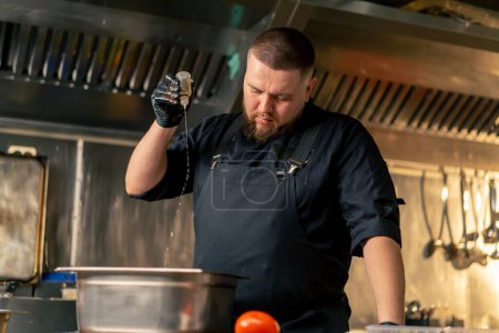 Foto de En la cocina profesional en un chef en una chaqueta negra vierte aceite en verduras en un recipiente de hierro - Imagen libre de derechos