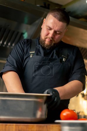 Téléchargez les photos : Dans une cuisine professionnelle en chef dans une veste noire mélange marinade et légumes dans un bol en fer - en image libre de droit