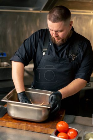 in einer professionellen Küche mixt Koch in schwarzer Jacke Marinade und Gemüse in einer eisernen Schüssel