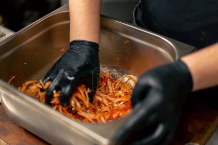 Téléchargez les photos : Close-up dans une cuisine professionnelle en chef dans une veste noire mélange marinade et légumes dans un bol en fer - en image libre de droit