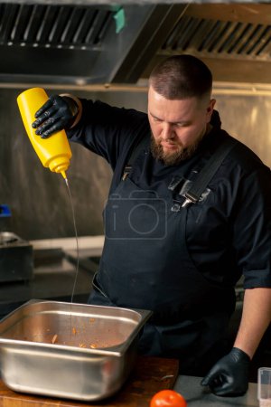 Téléchargez les photos : Dans la cuisine professionnelle dans un chef dans une veste noire verse de l'huile dans les légumes dans un bol de fer - en image libre de droit