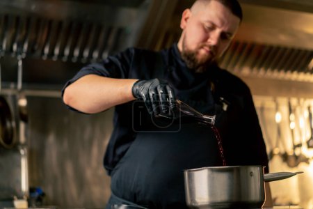 Téléchargez les photos : Dans la cuisine professionnelle un chef dans une veste noire verse de la sauce rouge dans une casserole debout sur la cuisinière - en image libre de droit