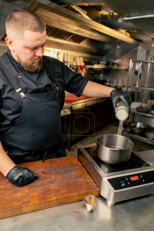 Téléchargez les photos : Dans un chef de cuisine professionnel dans une veste noire grésille sel dans une casserole debout sur la cuisinière - en image libre de droit