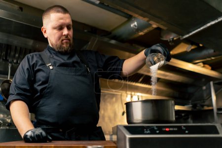 en una cocina profesional chef en una chaqueta negra chisporrotea sal en una cacerola de pie sobre la estufa