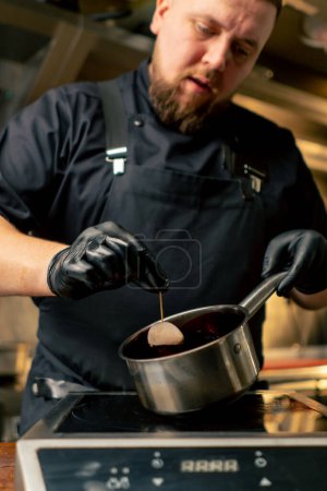 in einem professionellen Küchenchef in schwarzen Handschuhen tränkt eine Delikatesse in einer bordeauxroten Sauce aus der Pfanne