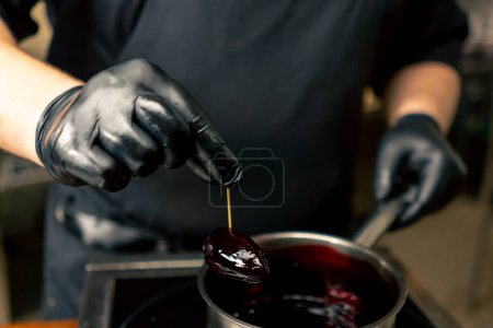 Téléchargez les photos : Gros plan dans une cuisine professionnelle chef en gants noirs imbibe une délicatesse dans une sauce bourgogne d'une casserole - en image libre de droit
