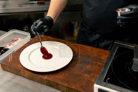 Foto de Primer plano en la cocina profesional con un chef que usa guantes negros salsa roja en una decoración de placa de cuchara - Imagen libre de derechos