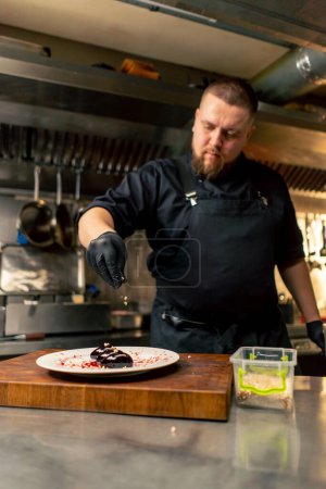 Photo for In a professional kitchen chef in a black jacket and gloves decorates a delicacy on a plate with nuts - Royalty Free Image