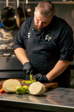 in a professional kitchen wearing black gloves cuts celery on wooden board