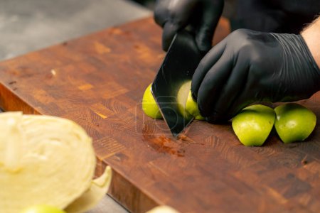 close-up in a professional kitchen wearing black gloves cuts a green apple on wooden board