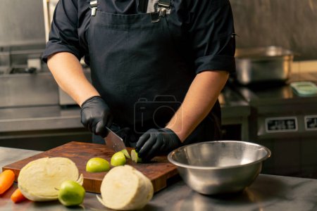 Foto de Primer plano en una cocina profesional con guantes negros corta una manzana verde en el tablero de madera - Imagen libre de derechos