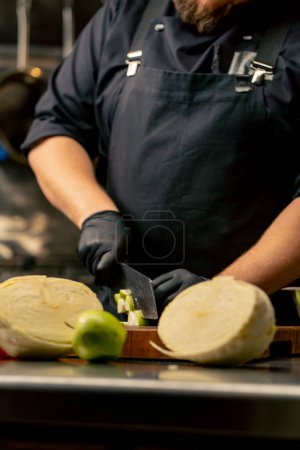 Téléchargez les photos : Gros plan dans une cuisine professionnelle portant des gants noirs coupe une pomme verte sur une planche de bois - en image libre de droit