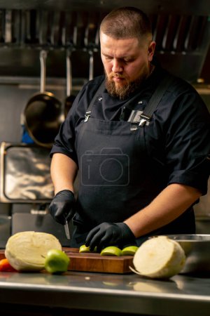 in a professional kitchen wearing black gloves cuts a green apple on wooden board