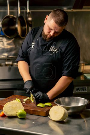 Foto de En una cocina profesional con guantes negros corta una manzana verde en una tabla de madera - Imagen libre de derechos
