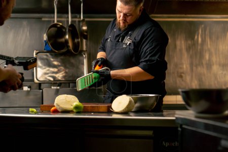 Foto de En una cocina profesional con guantes negros corta zanahorias en un rallador verde - Imagen libre de derechos