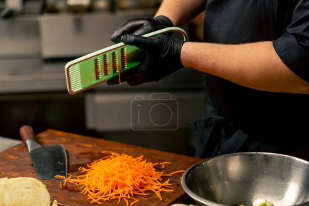 Nahaufnahme in einer professionellen Küche mit schwarzen Handschuhen schneidet Möhren auf einer grünen Reibe