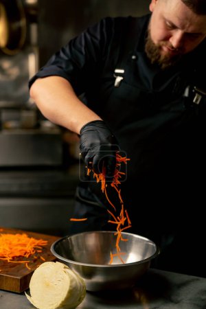 Photo for In a professional kitchen wearing black gloves pours carrot sticks into an iron bowl - Royalty Free Image