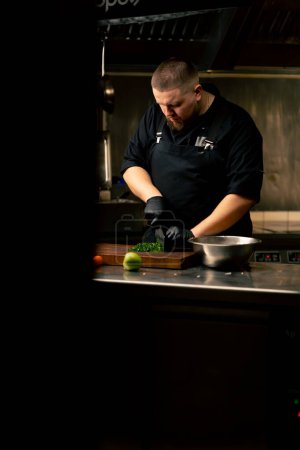 in einer professionellen Küche schneidet ein Koch in schwarzer Jacke und Handschuhen Petersilie auf dem Tisch