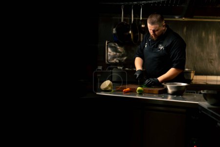 Photo for In a professional kitchen a chef in a black jacket and gloves chops parsley on the table - Royalty Free Image