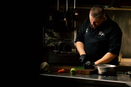 in a professional kitchen a chef in a black jacket and gloves chops parsley on the table