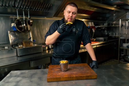 Photo for In a professional kitchen a chef in a black jacket tastes freshly prepared French fries - Royalty Free Image