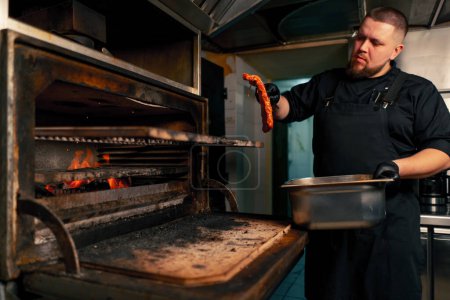 Téléchargez les photos : Dans une cuisine professionnelle, un chef vêtu d'une veste noire dispose des côtes marinées près d'un four à grill chaud - en image libre de droit