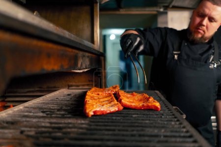 Photo for In a professional kitchen a chef in a black jacket lays out marinated ribs near a hot grill oven - Royalty Free Image