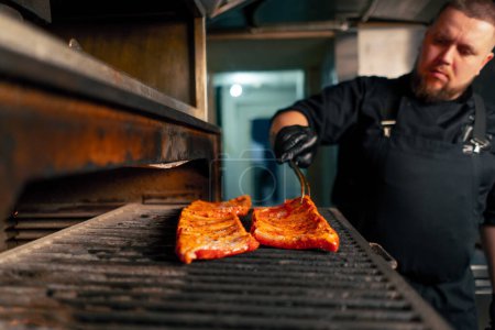 Photo for In a professional kitchen a chef in a black jacket lays out marinated ribs near a hot grill oven - Royalty Free Image